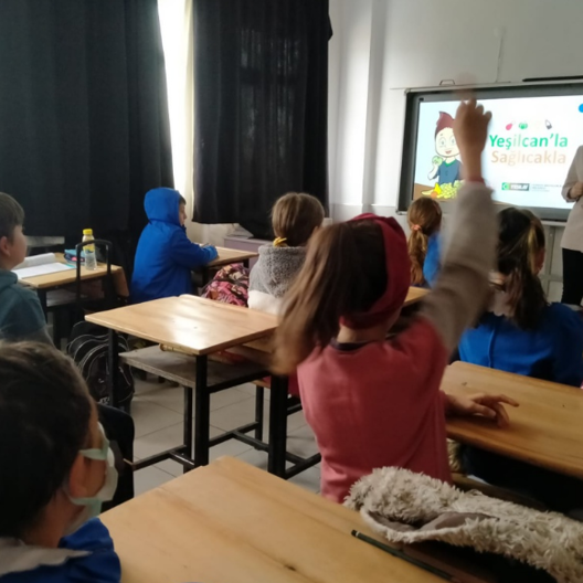 Bir sınıfın sağ arka köşesinden çekilmiş fotoğrafta, tek kişilik sıralarda oturan önlüklü ve sivil öğrenciler, karşılarındaki akıllı tahtaya bakıyor. Orta sıralarda oturan sivil giyimli bir kız öğrenci parmak kaldırmış.  Tahtadaki çizimde kahverengi saçlı bir oğlan çocuğu masaya oturmuş. Önündeki tabakta çeşitli meyveler var. Yanında,  “Yeşilcan’la Sağlıcakla” yazıyor. 