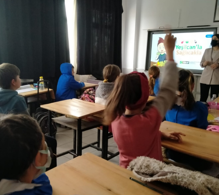 Bir sınıfın sağ arka köşesinden çekilmiş fotoğrafta, tek kişilik sıralarda oturan önlüklü ve sivil öğrenciler, karşılarındaki akıllı tahtaya bakıyor. Orta sıralarda oturan sivil giyimli bir kız öğrenci parmak kaldırmış.  Tahtadaki çizimde kahverengi saçlı bir oğlan çocuğu masaya oturmuş. Önündeki tabakta çeşitli meyveler var. Yanında,  “Yeşilcan’la Sağlıcakla” yazıyor. 