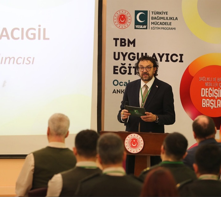 Fotoğrafta kıvırcık saçlı, gözlüklü, kısa sakallı, kumral bir adam, Türkiye Cumhuriyeti Millî Savunma Bakanlığı logolu kürsüye geçmiş, ellerinin arasındaki Yeşilay logolu kartla, salondaki askeri üniformalı katılımcılara eğitim veriyor. Arkasındaki fonun sol üst köşesinde Türkiye Cumhuriyeti Millî Savunma Bakanlığı’nın logosu, Yeşilay’ın logosu ve Türkiye Bağımlılıkla Mücadele Eğitim Programı” yazısı var. Logoların altında “TBM UYGULAYICI EĞİTİM” yazısı yer almış.